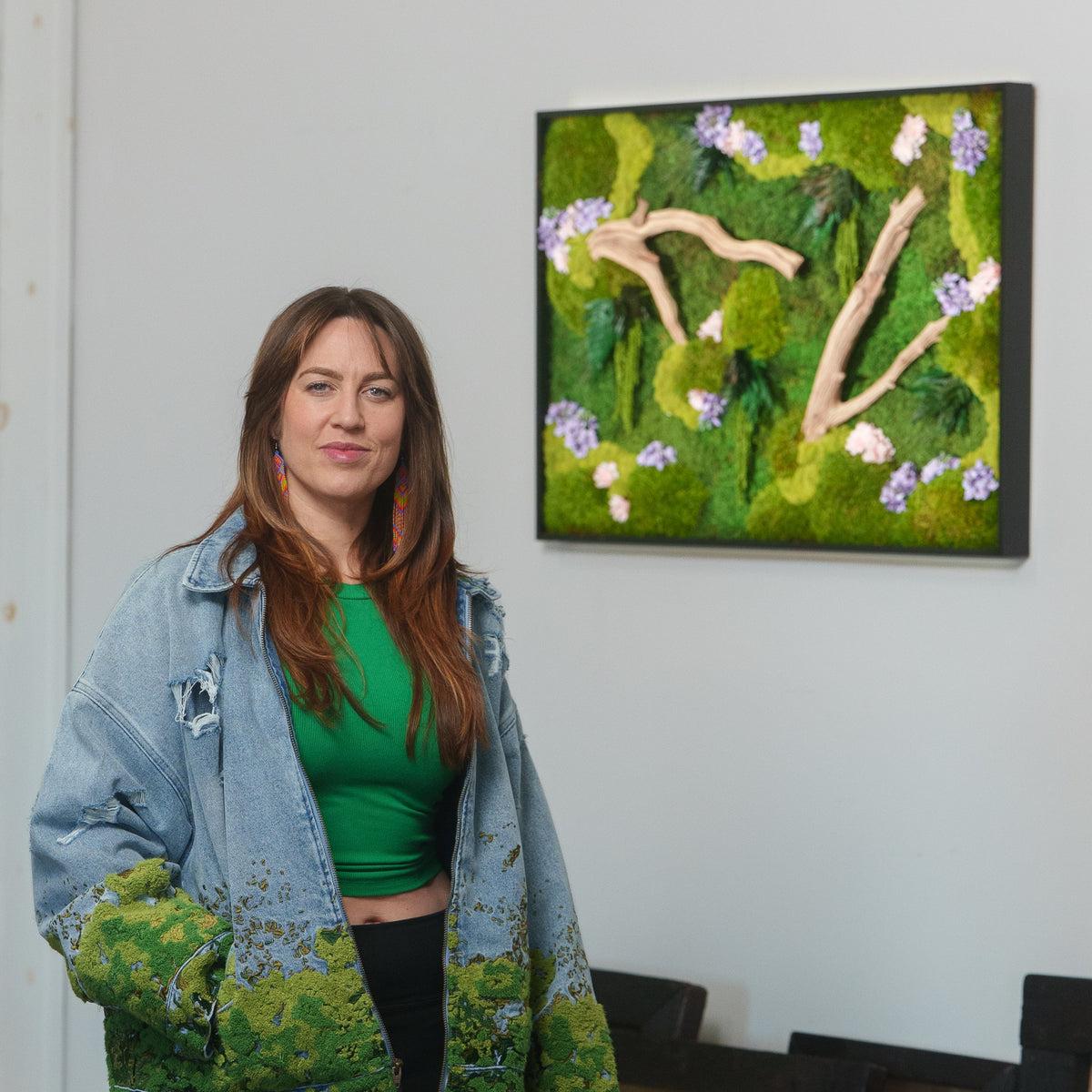 Kate the Earthling standing beside a framed preserved moss piece with driftwood branches and purple and pink flowers, wearing a denim jacket covered in moss