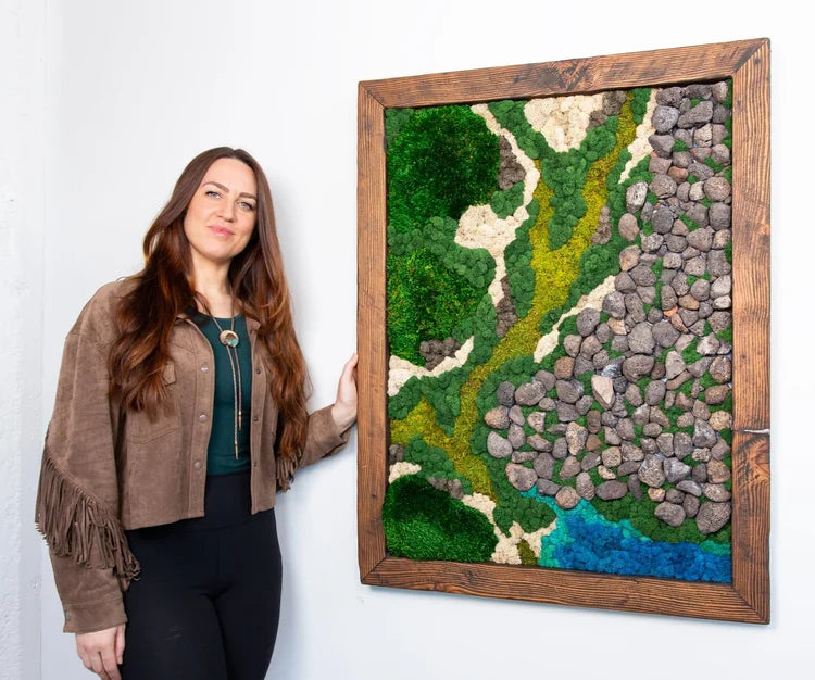 Kate Shooltz standing beside Akela the Rock Glacier, a framed moss artwork with stone scree and blue meltwater