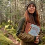 Kate Shooltz holding her children's book Kate the Earthling on a mossy forest trail
