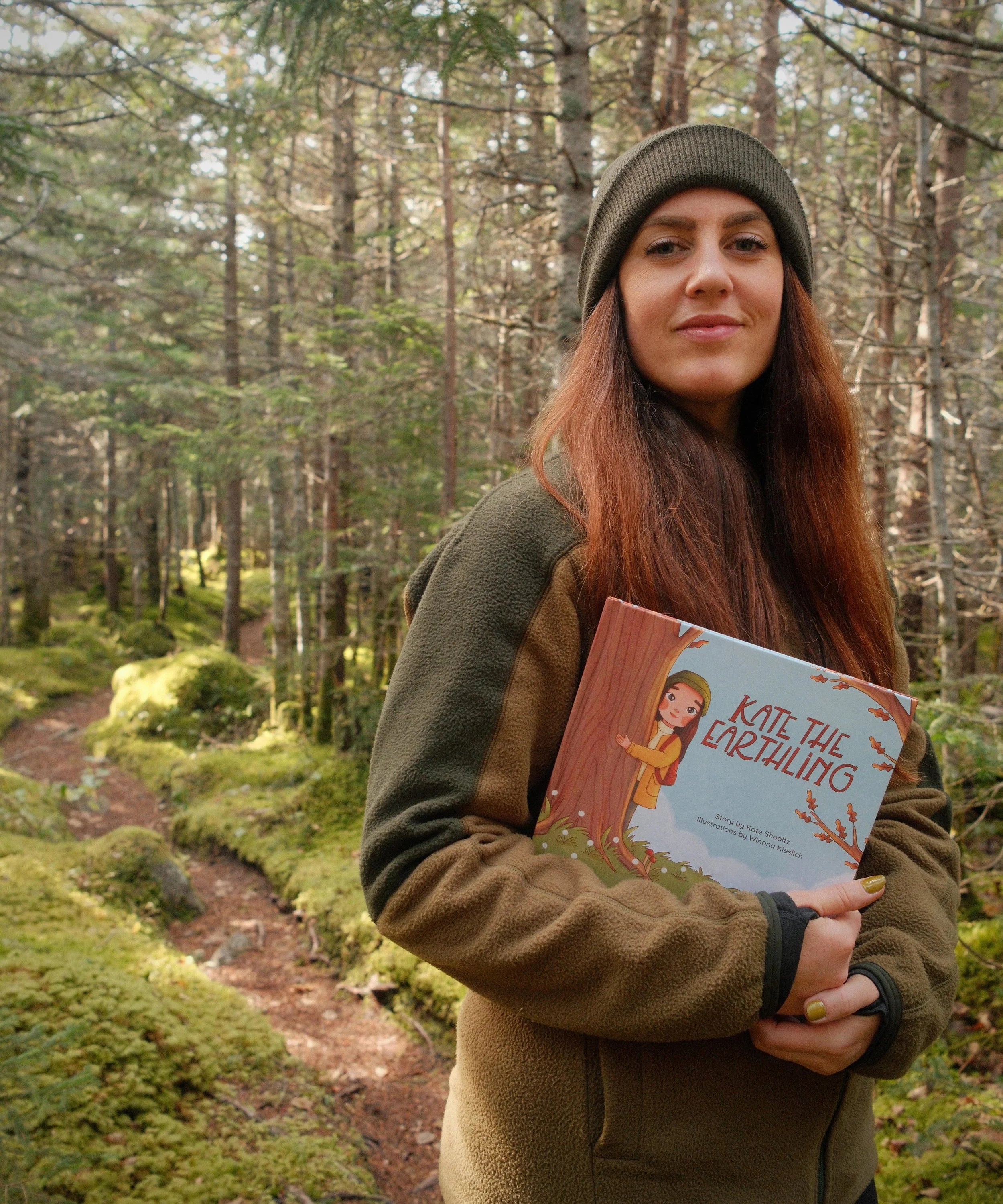 Kate Shooltz holding her children's book Kate the Earthling on a mossy forest trail
