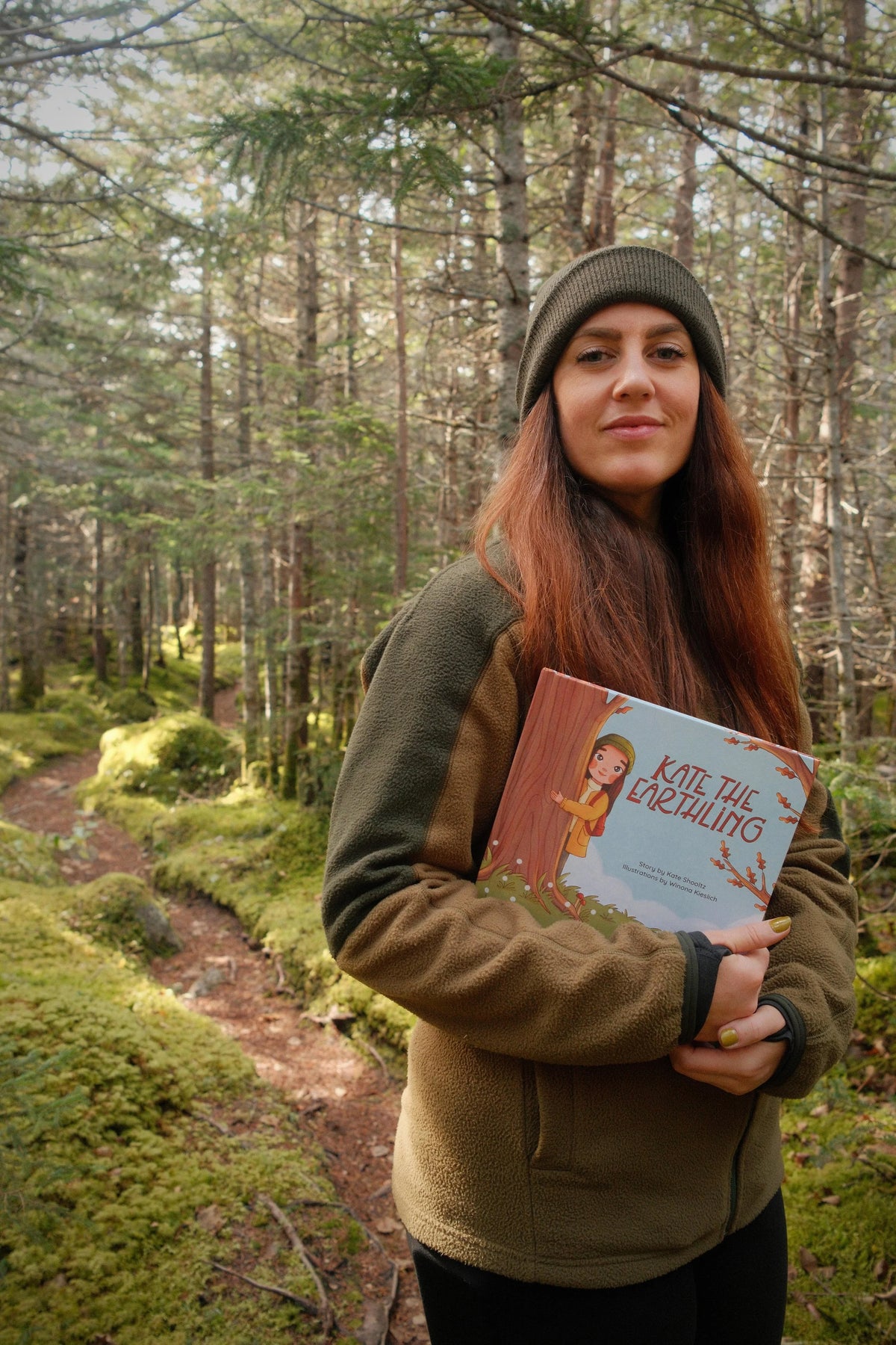Kate Shooltz holding her children's book Kate the Earthling on a mossy forest trail