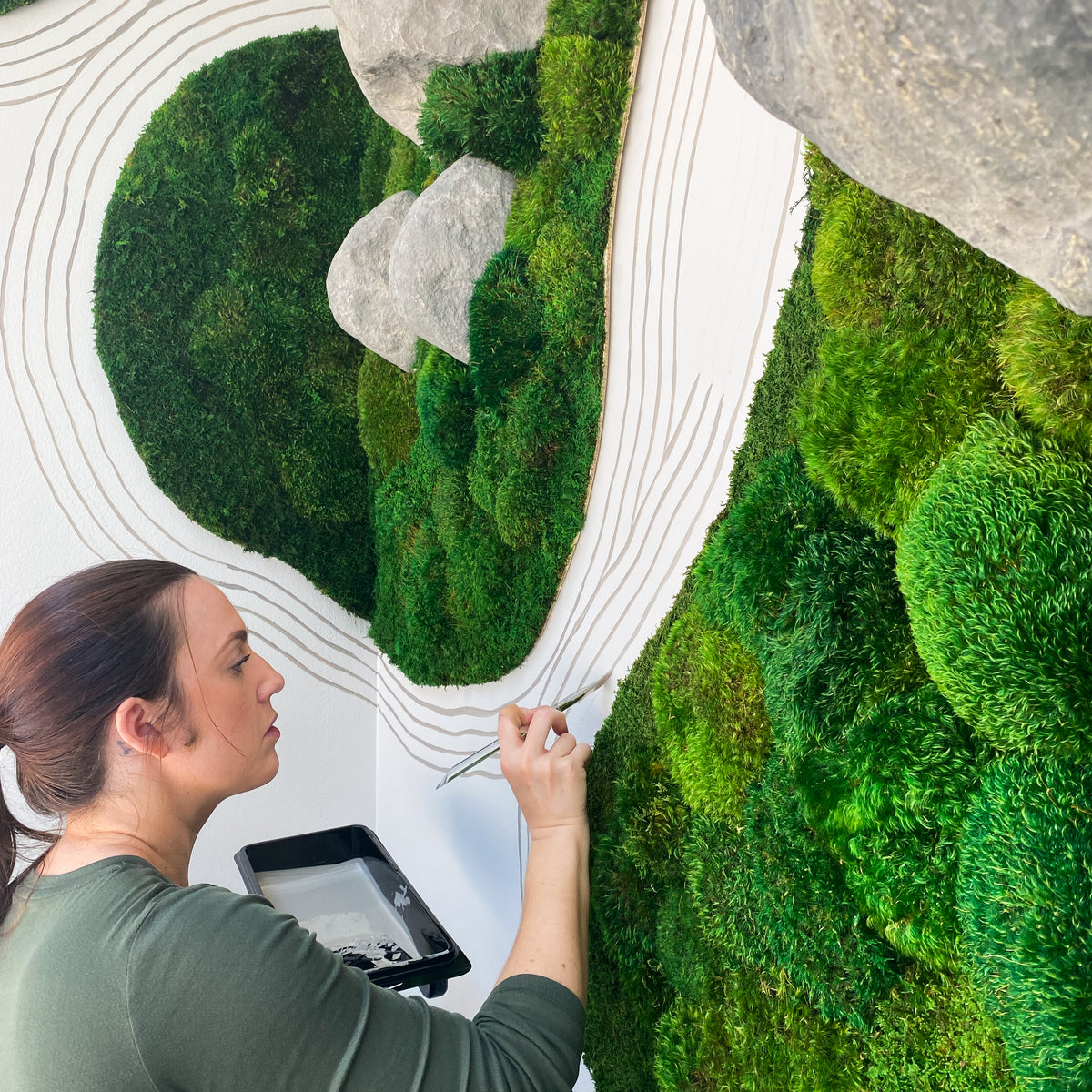 Kate Shooltz working on a large moss wall with carved white contour lines and stone forms