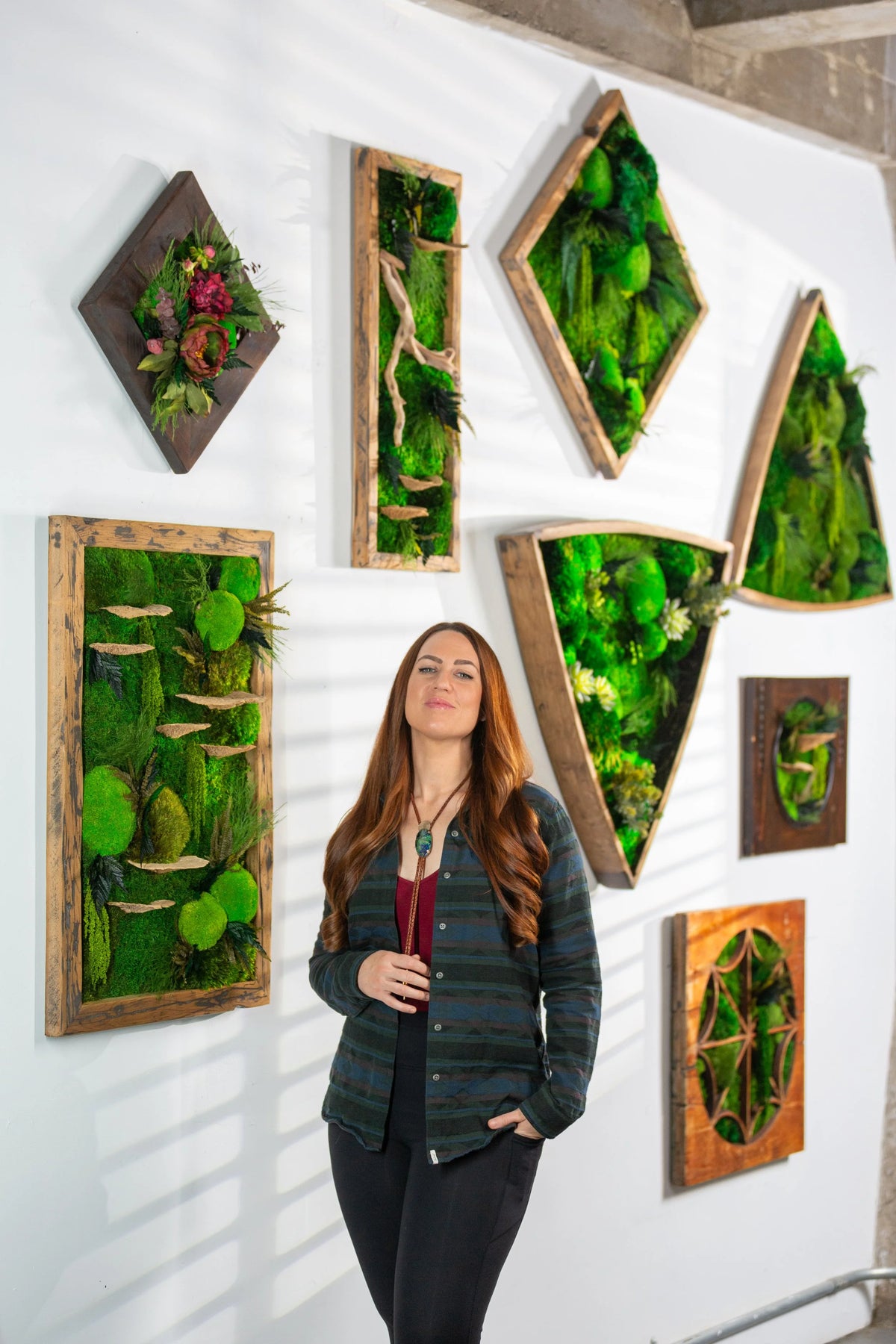 Kate Shooltz seated below a wall of angular framed moss panels with winding pale paths
