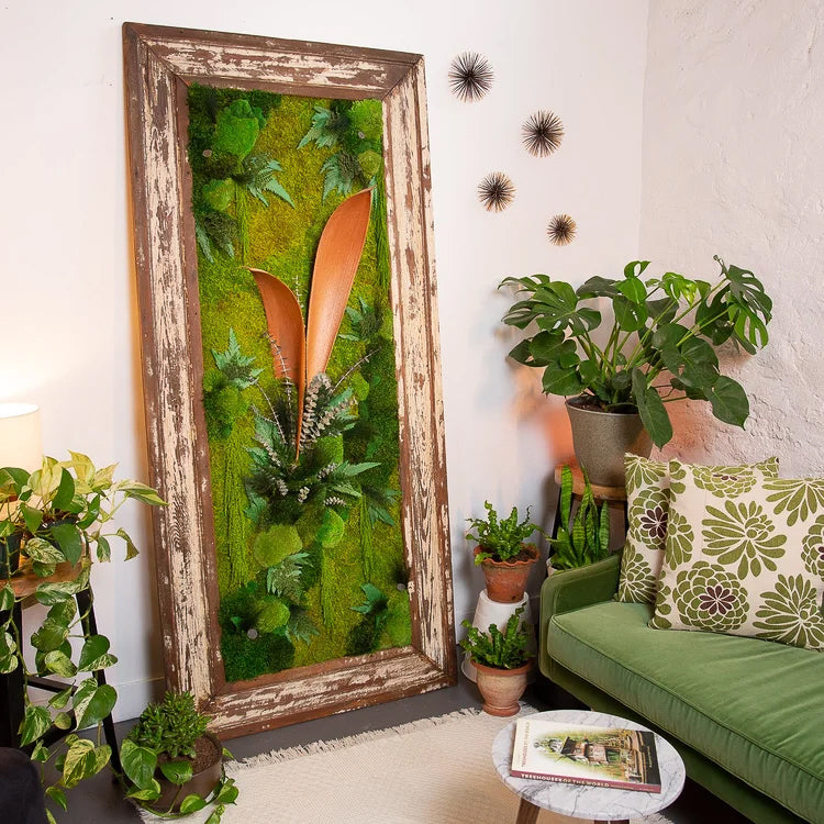 Tall distressed white frame filled with moss and orange leaf forms, beside a green sofa and potted plants