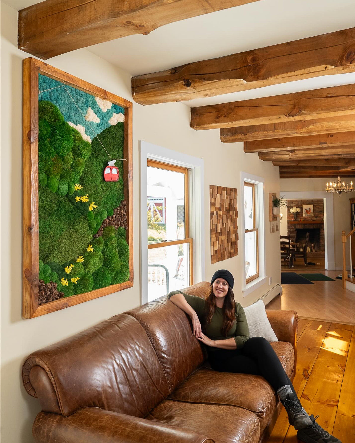 Wood framed moss artwork of a mountain cable car scene on a beamed ceiling wall above a leather sofa