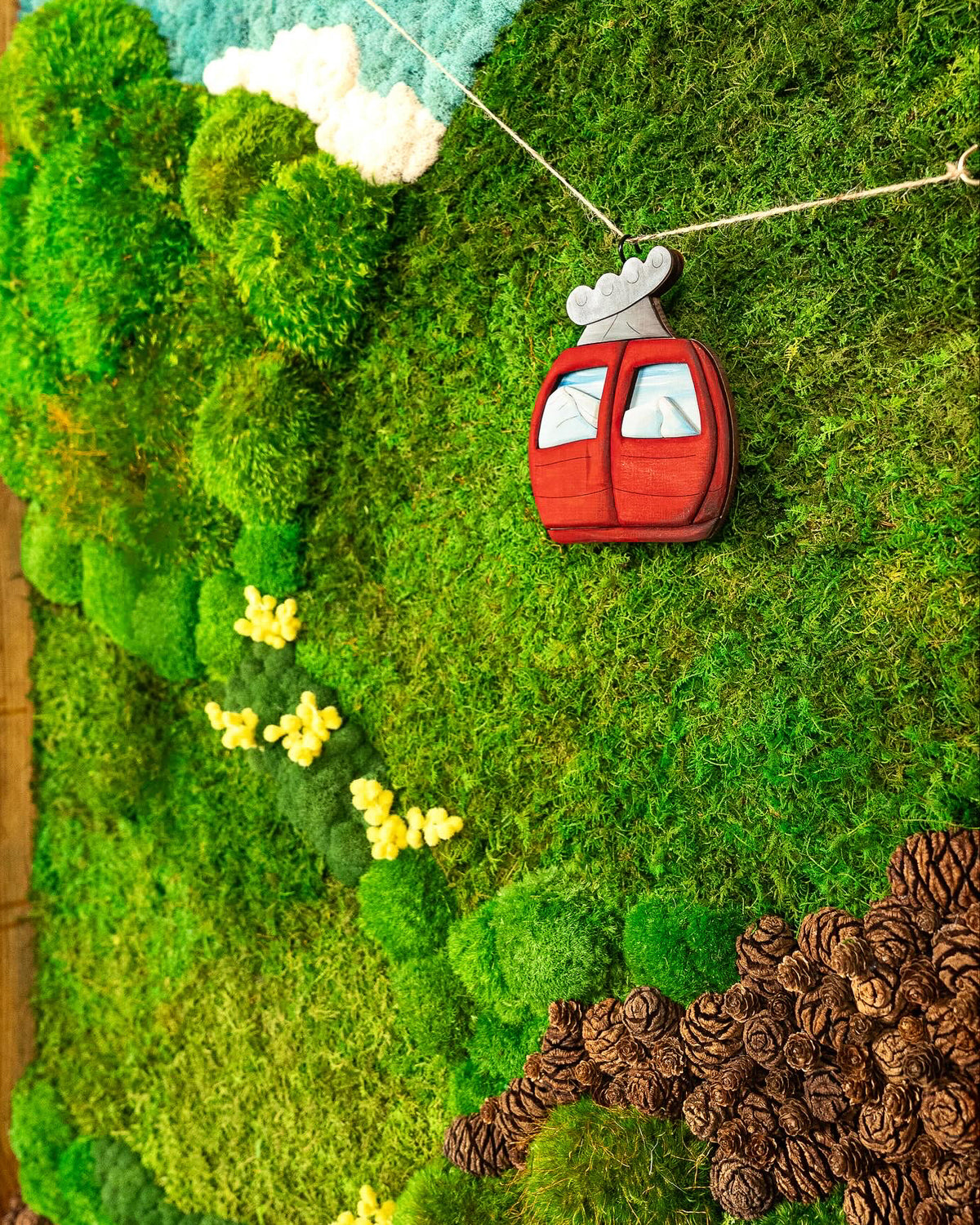 Close up of a moss landscape with a red cable car, blue sky, white clouds, yellow blooms and pinecones