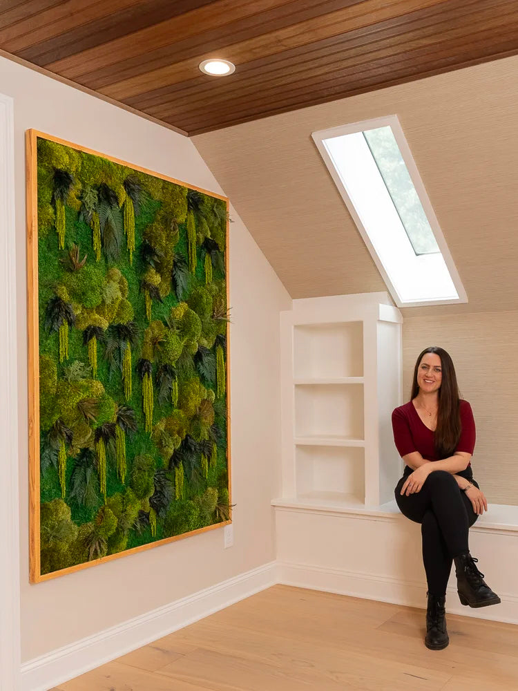 Large wood framed fern and moss wall panel under a sloped plank ceiling, with the artist seated on the bench beside it
