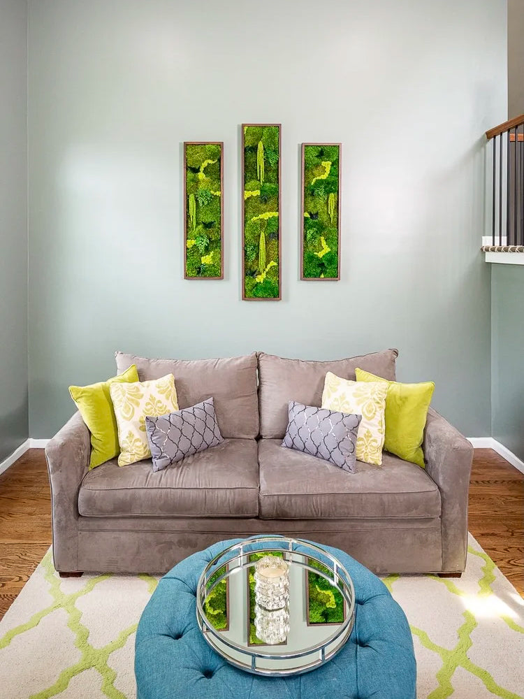Three narrow vertical moss panels on a pale green wall above a taupe sofa with yellow and purple pillows