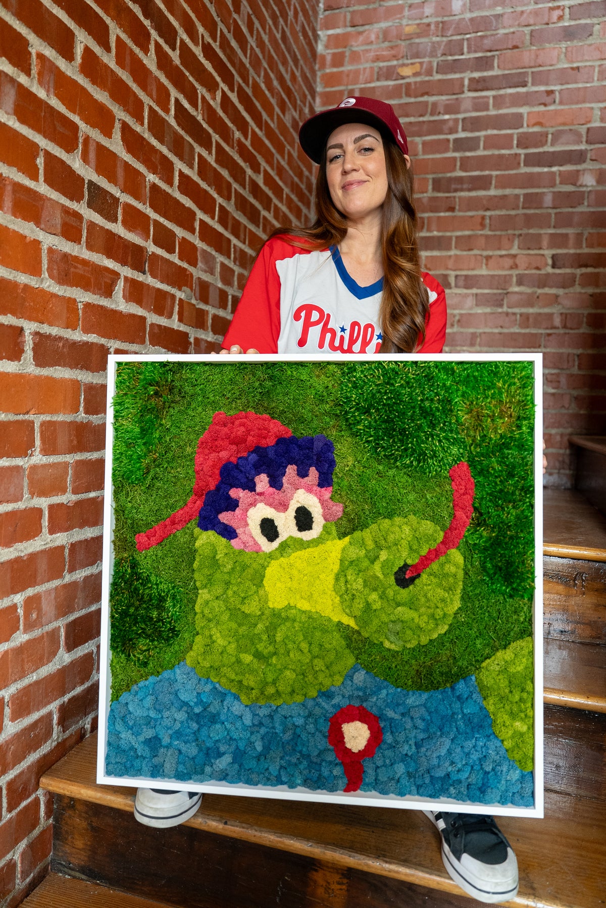 The artist in a Phillies jersey holding the preserved moss Phillie Phanatic portrait on a staircase